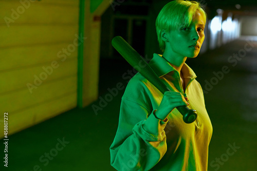 a teenage girl with short hair holding a baseball bat and looking straight into the camera. Modern parking in neon green light. High quality photo