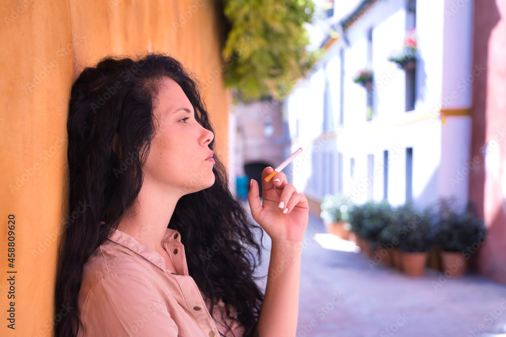 Middle-aged adult Hispanic woman with black curly hair, leaning against a yellow wall smoking. Concept smoking, cigar, tobacco, health.