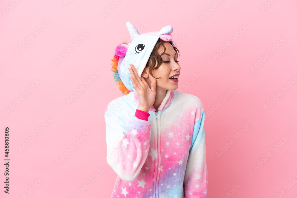 Young caucasian woman with unicorn pajamas isolated on pink background listening to something by putting hand on the ear