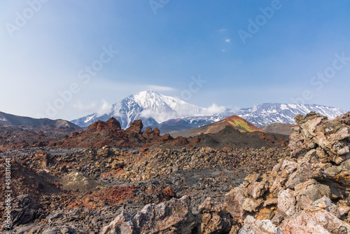 Red volcanic rocks against the background of Kamchatka volcanoes with snow-capped peaks in the clouds.