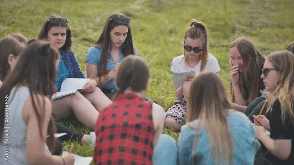 A group of female students are sitting in a circle on a meadow for collective work with notebooks. A group of female students are sitting in a circle on a meadow for collective work with notebooks.