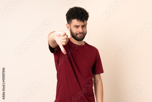 Young Moroccan man isolated on beige background showing thumb down with negative expression