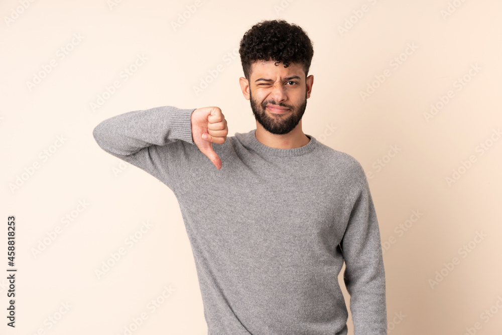 Young Moroccan man isolated on beige background showing thumb down with negative expression Young Moroccan man isolated on beige background showing thumb down with negative expression