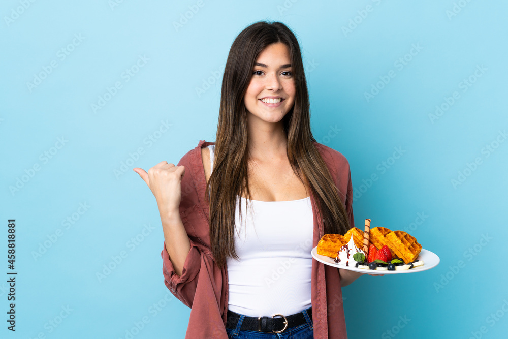 Teenager Brazilian girl holding waffles over isolated blue background pointing to the side to present a product Teenager Brazilian girl holding waffles over isolated blue background pointing to the side to present a product