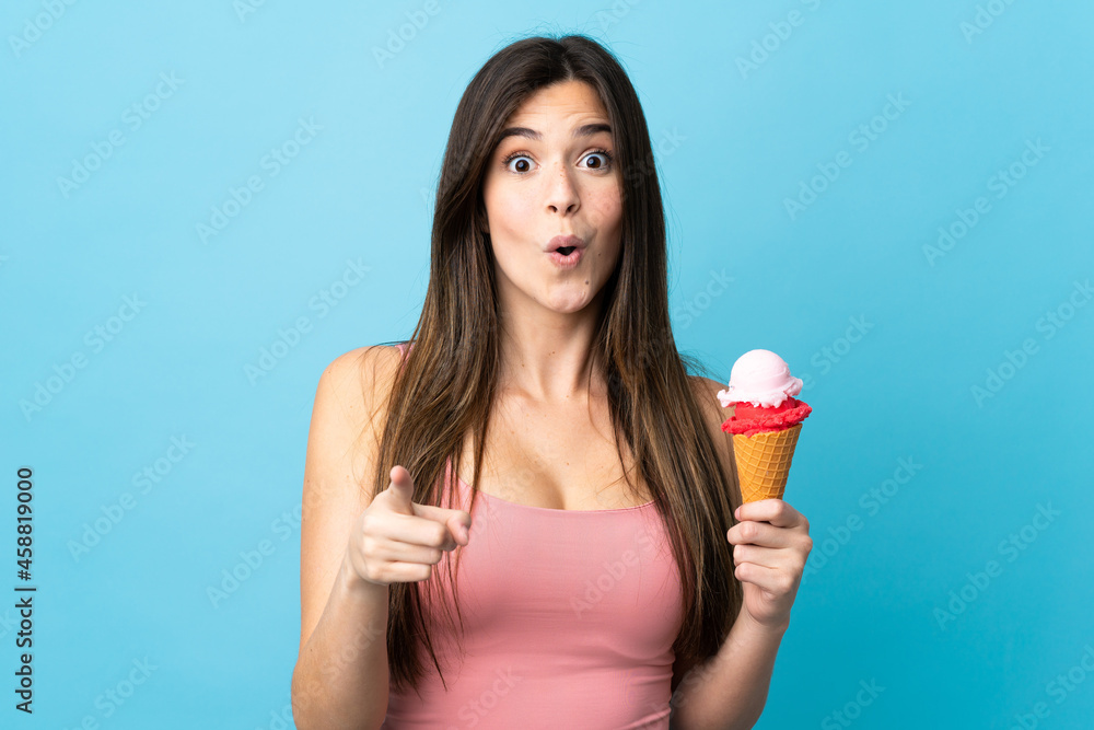 Teenager Brazilian girl holding a cornet ice cream over isolated blue background surprised and pointing front Teenager Brazilian girl holding a cornet ice cream over isolated blue background surprised and pointing front