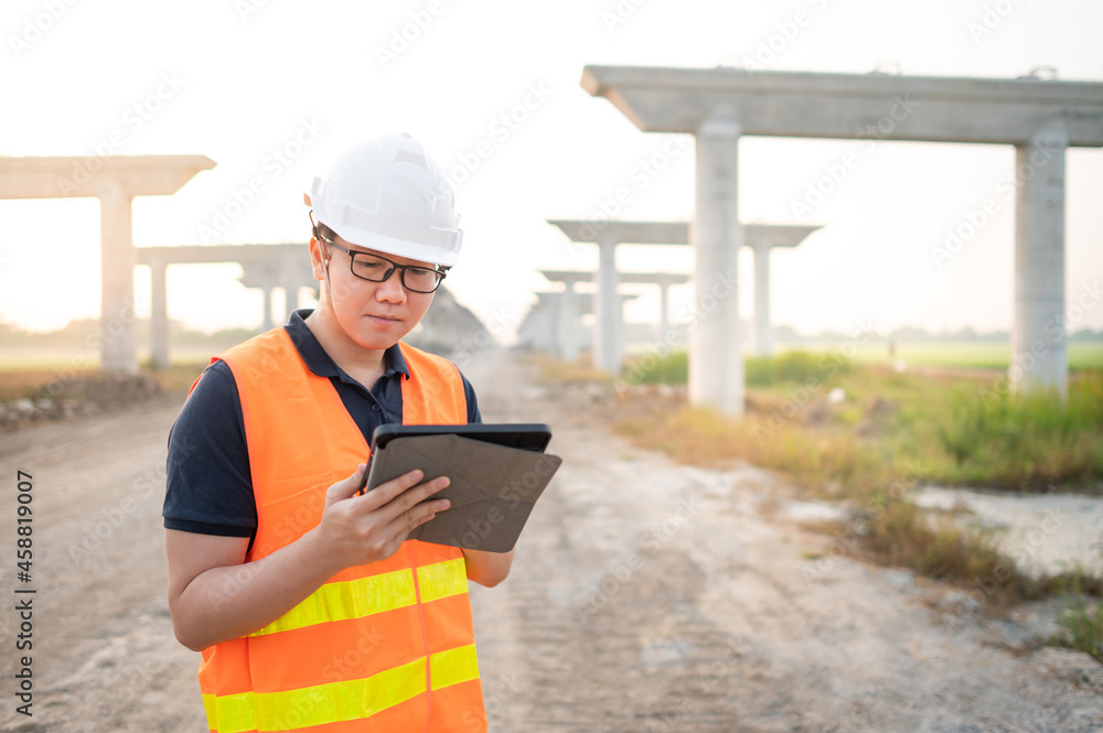 Smart Asian worker man or male civil engineer with protective safety helmet and reflective vest using digital tablet for project planning and checking architectural drawing at construction site. Smart Asian worker man or male civil engineer with protective safety helmet and reflective vest using digital tablet for project planning and checking architectural drawing at construction site.