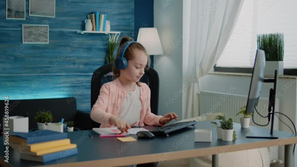 Young child drawing on textbook and using computer while wearing headphones for remote online class lesson. Schoolgirl using colorful pencils to draw on notebook and typing on keyboard. Young child drawing on textbook and using computer while wearing headphones for remote online class lesson. Schoolgirl using colorful pencils to draw on notebook and typing on keyboard.