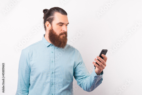 Serious bearded man is looking at the phone he is holding over white background. © Vulp