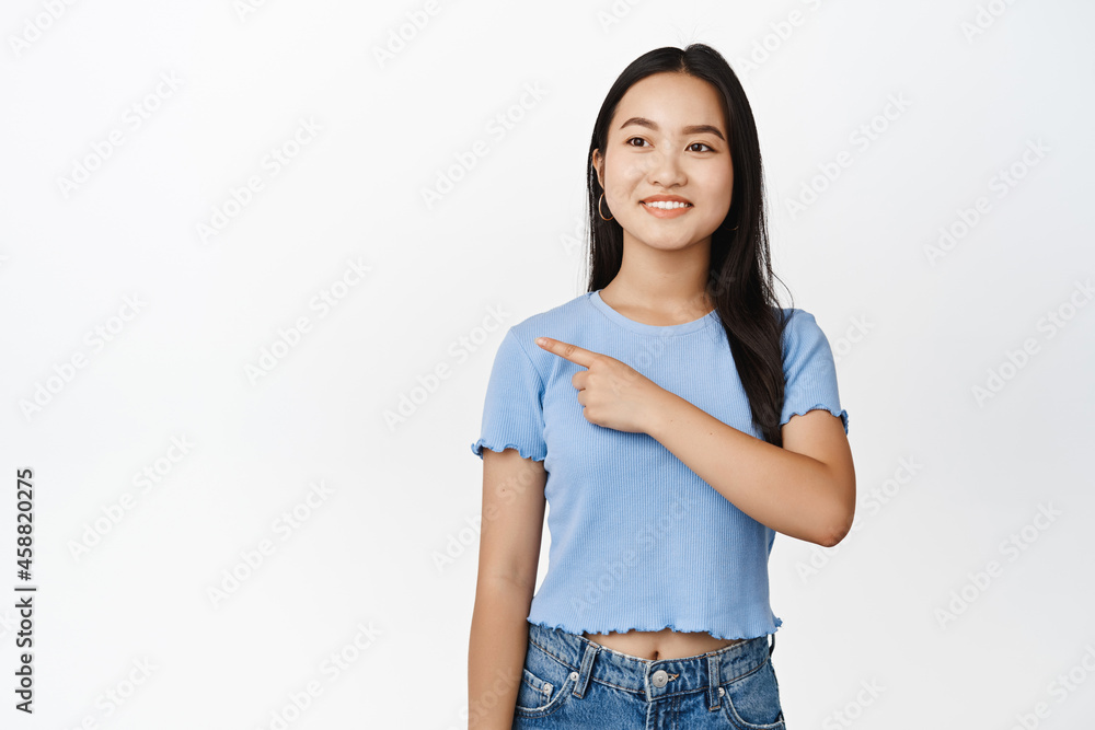 Attractive brunette asian woman pointing at upper left corner, smiling and gazing at promo offer, standing in tshirt over white background Attractive brunette asian woman pointing at upper left corner, smiling and gazing at promo offer, standing in tshirt over white background