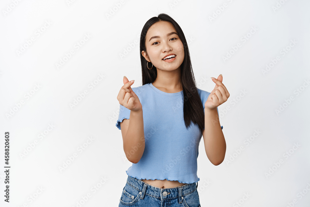 Cute brunette asian girl showing finger hearts and smiling, like something, standing in tshirt over white background Cute brunette asian girl showing finger hearts and smiling, like something, standing in tshirt over white background