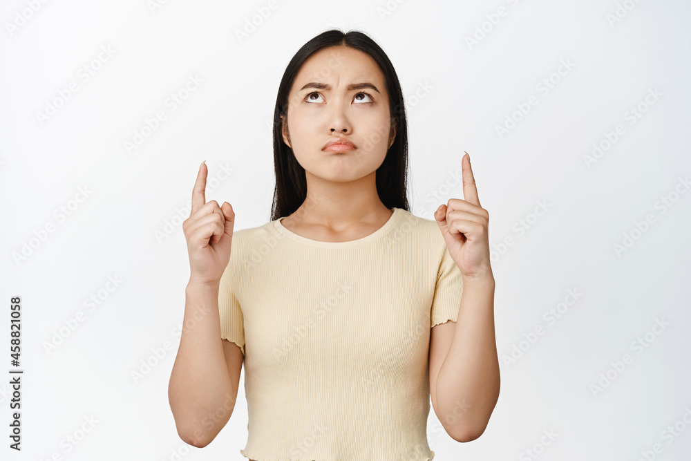 Upset sulking asian woman pointing and looking up with disappointed, jealous face, standing in yellow t-shirt over white background Upset sulking asian woman pointing and looking up with disappointed, jealous face, standing in yellow t-shirt over white background