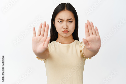 Close up portrait of serious asian girl, stretch out hands in stop gesture, prohibit something, forbid, refusing smth bad, standing over white background © Cookie Studio