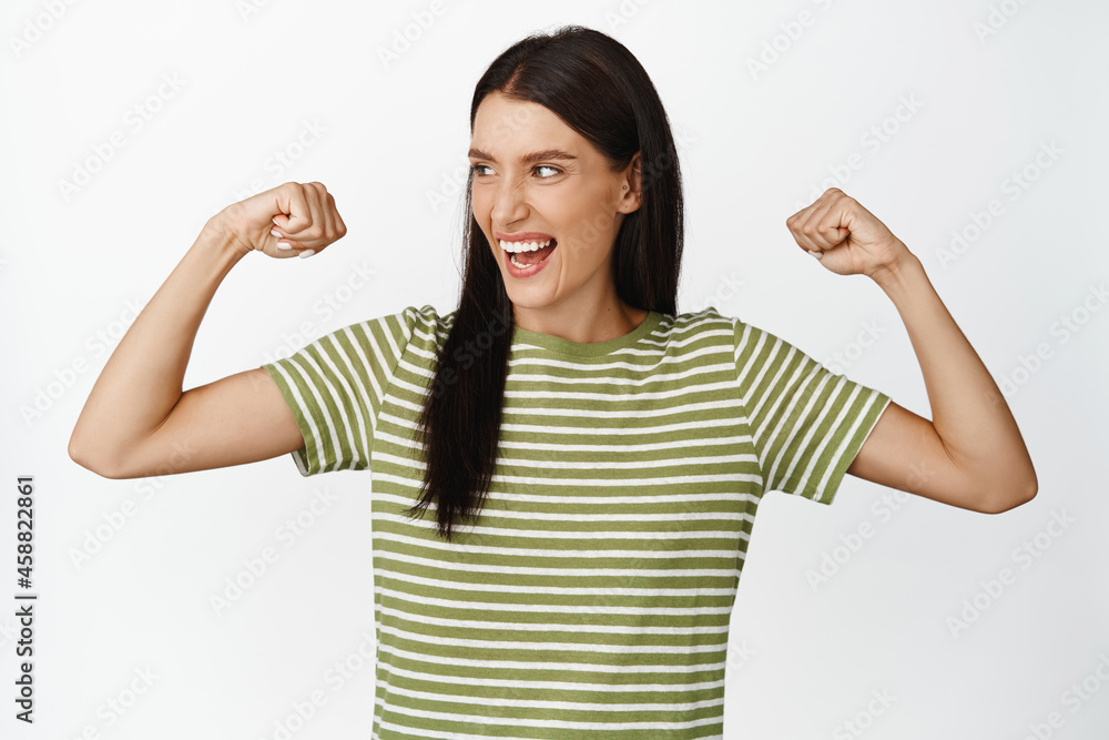 Empowered happy woman feeling strong, flexing biceps, showing her muscles on arms and laughing, standing over white background Empowered happy woman feeling strong, flexing biceps, showing her muscles on arms and laughing, standing over white background