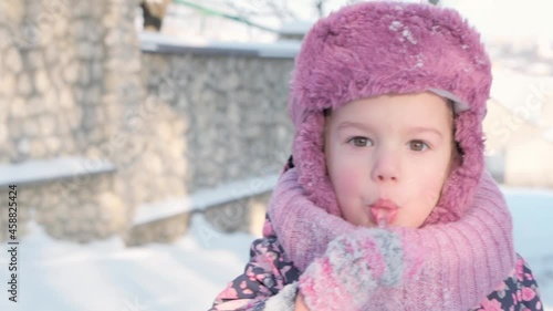Winter, vacation, games, family concepts - slo-mo close up of authentic little preschool minor 3-4 years old girl in purple looking at camera licks icicle with tongue on snow. child runs on snowy area photo