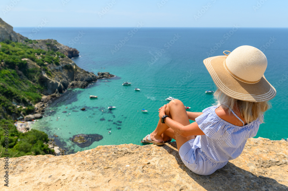 A young girl in a straw hat sits on the edge of a cliff and looks at the beautiful seascape with yachts and a transparent sea. Travel across the Crimea.