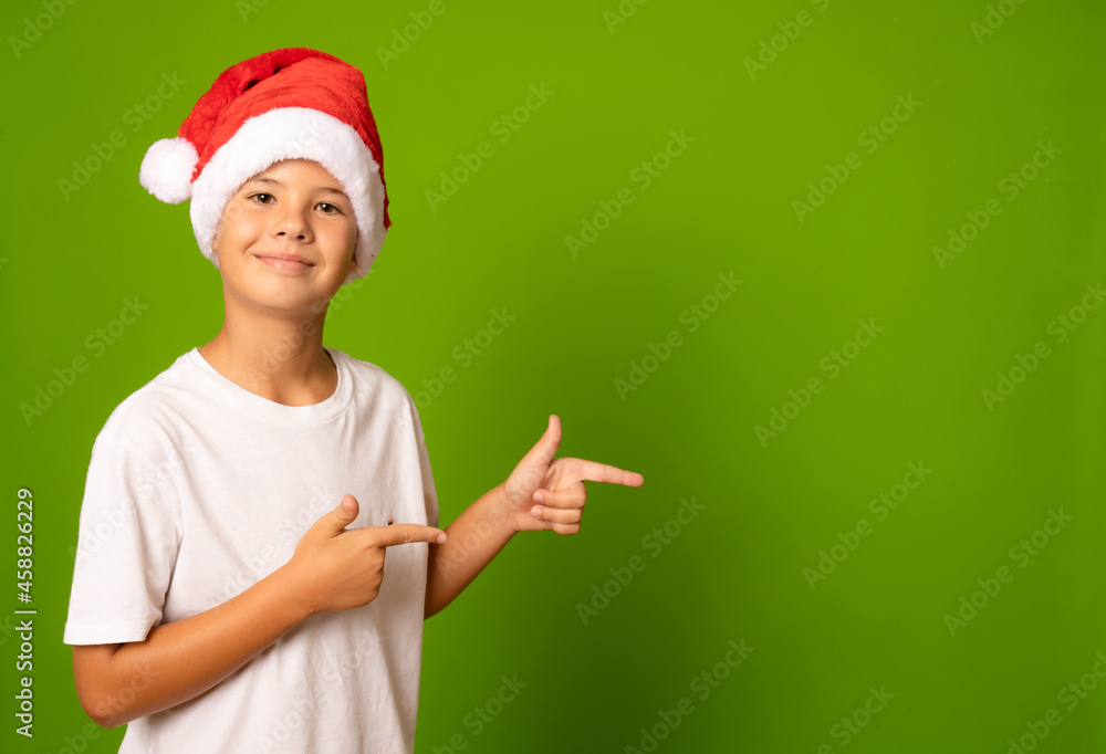 Portrait of a beautiful smiling boy in a red hat and white t-shirt pointing fingers side on a green background. A child in a Christmas hat of Santa Claus. Portrait of a beautiful smiling boy in a red hat and white t-shirt pointing fingers side on a green background. A child in a Christmas hat of Santa Claus.