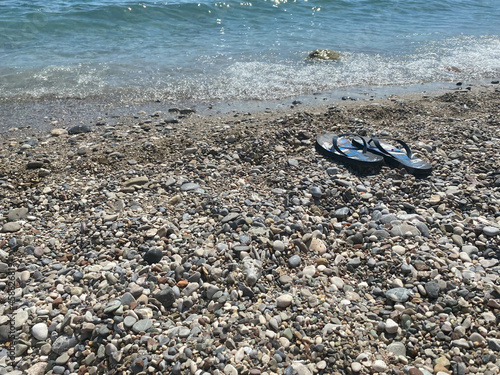 Blue flip-flops on the pebble beach with turquoise sea and blue sky in background. Summertime. Vacation concept