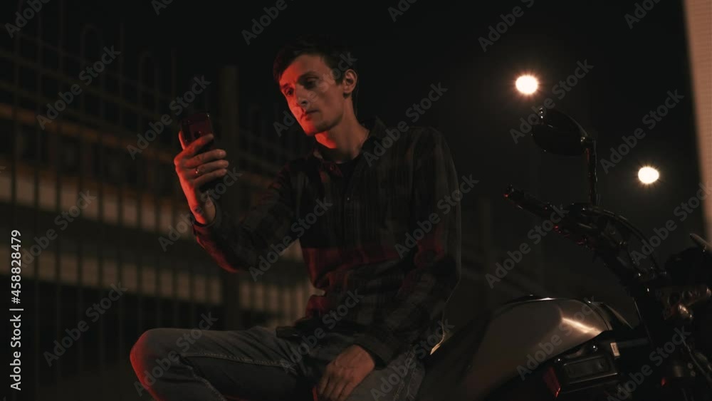 Portrait of young attractive man motorcyclist on night street in red light using smartphone Portrait of young attractive man motorcyclist on night street in red light using smartphone