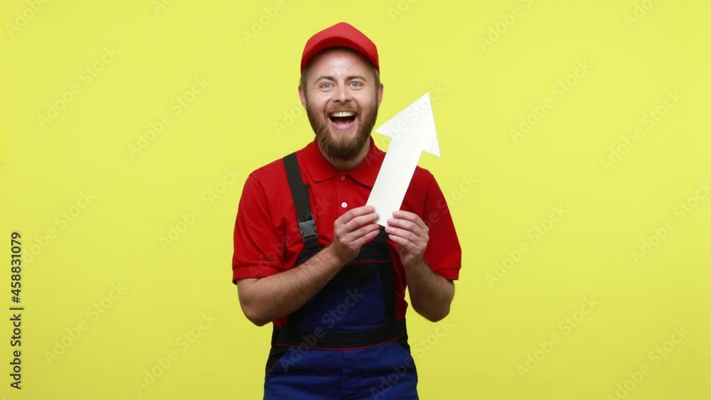 Extremely happy worker showing white paper arrow pointing up, showing increase of orders and salary, wearing blue uniform, red T-shirt and visor cap. Indoor studio shot isolated over yellow background Extremely happy worker showing white paper arrow pointing up, showing increase of orders and salary, wearing blue uniform, red T-shirt and visor cap. Indoor studio shot isolated over yellow background
