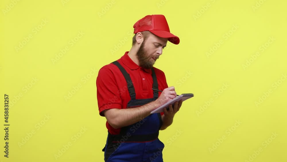 Pensive handyman standing with paper organizer, making working plan or to do list, wearing blue uniform, red T-shirt and visor cap. Indoor studio shot isolated over yellow background. Pensive handyman standing with paper organizer, making working plan or to do list, wearing blue uniform, red T-shirt and visor cap. Indoor studio shot isolated over yellow background.