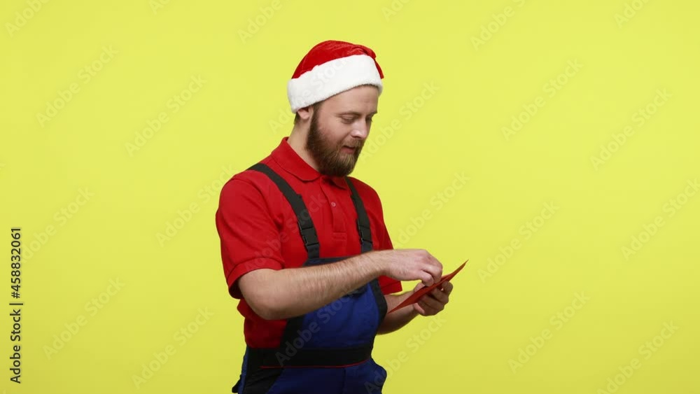 Side view of touched worker holding red envelope and reading letter from mysterious stranger, wearing blue overalls and santa claus hat. Indoor studio shot isolated over yellow background. Side view of touched worker holding red envelope and reading letter from mysterious stranger, wearing blue overalls and santa claus hat. Indoor studio shot isolated over yellow background.