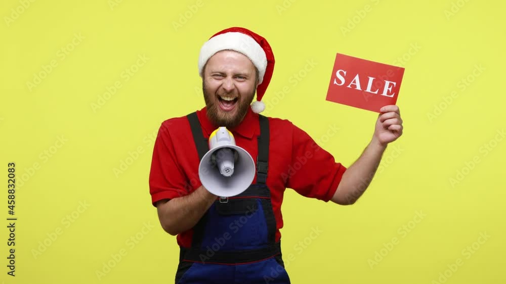 Shop assistant screaming in megaphone and showing card with sale inscription, has excited look, wearing blue overalls and santa claus hat. Indoor studio shot isolated over yellow background. Shop assistant screaming in megaphone and showing card with sale inscription, has excited look, wearing blue overalls and santa claus hat. Indoor studio shot isolated over yellow background.