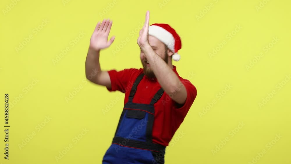 Extremely happy optimistic worker dancing on corporate Christmas party, being in excellent mood, wearing blue overalls and santa claus hat. Indoor studio shot isolated over yellow background. Extremely happy optimistic worker dancing on corporate Christmas party, being in excellent mood, wearing blue overalls and santa claus hat. Indoor studio shot isolated over yellow background.