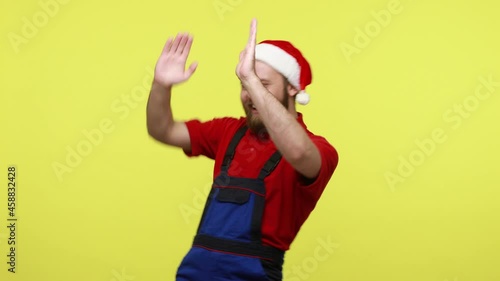 Extremely happy optimistic worker dancing on corporate Christmas party, being in excellent mood, wearing blue overalls and santa claus hat. Indoor studio shot isolated over yellow background.