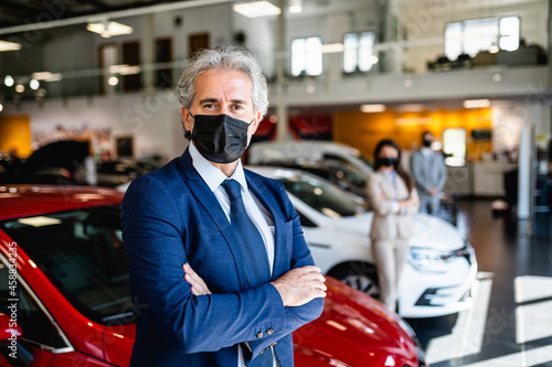 Senior salesman with protective face mask working at car showroom. Customers in the background.