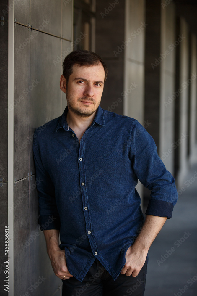 Young man weared in blue denim shirt on background of the City. Fashion style guy classic portrait. Street style male portrait looking to camera. Contemporary businessman. Modern urban walk in a town Young man weared in blue denim shirt on background of the City. Fashion style guy classic portrait. Street style male portrait looking to camera. Contemporary businessman. Modern urban walk in a town