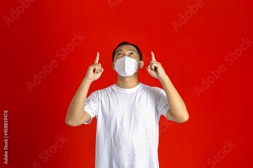 Asian man wearing a medical mask pointing up with both hands while looking up on a red background