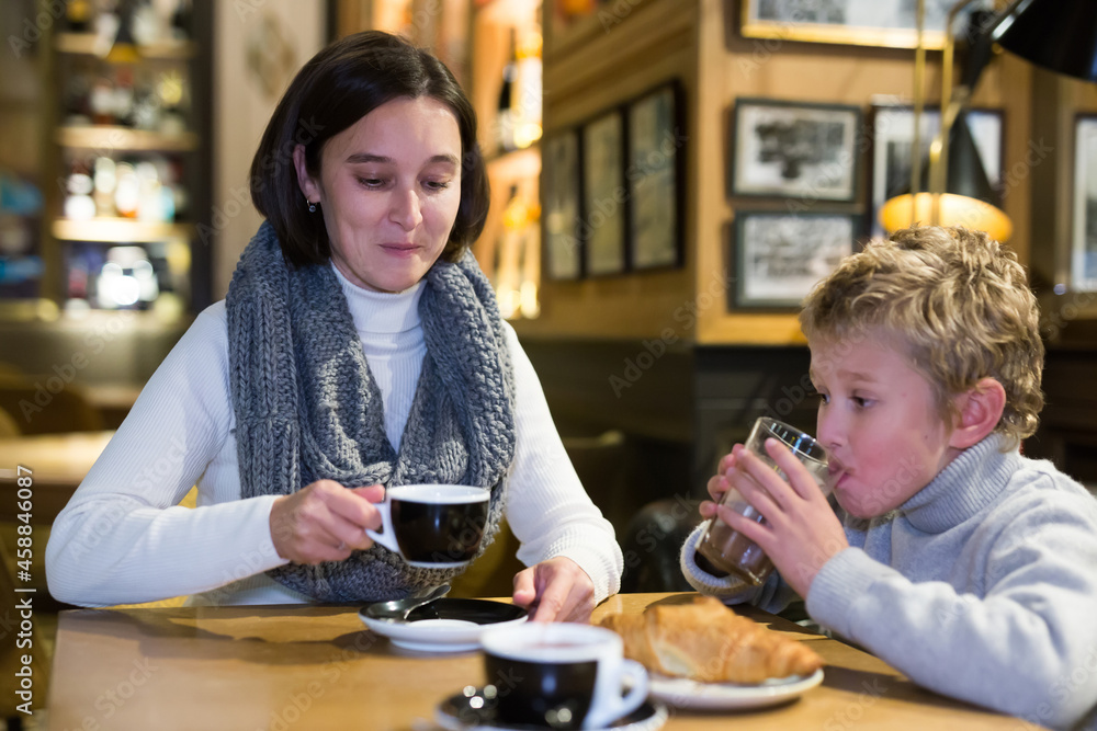 Young mother with tween son having fun at cafe, drinking coffee and cocoa with croissants and friendly talking