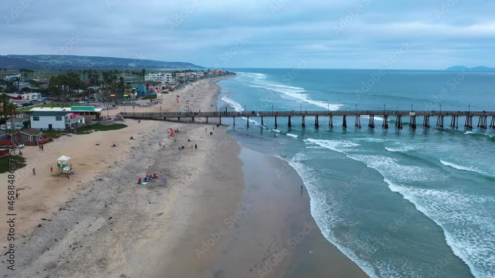 Drone flies low over beach and tourists walking in the sand towards pier Drone flies low over beach and tourists walking in the sand towards pier