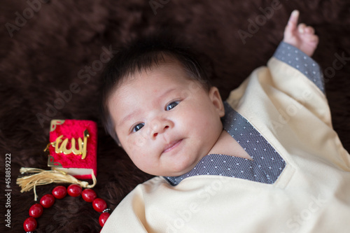 A cute Asian infant Muslim in thobe and rosary beads on red, muslim bead on bed in the home, translation is the name of muslim God 