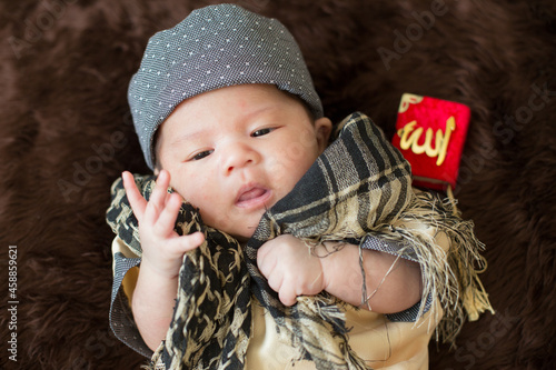 A cute Asian infant Muslim in thobe with prayer cap on bed in home, translation is noble quran photo