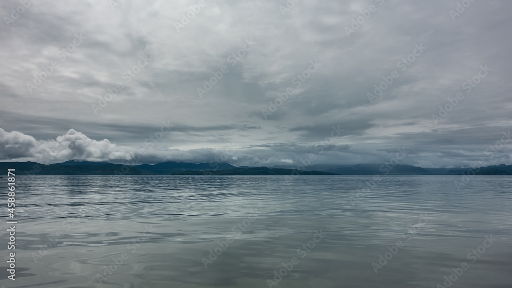 The ocean under a cloudy sky. Reflection on a shiny surface. In the distance, you can see a mountain range, the peaks of which are hidden in the clouds. Pastel shades. Petropavlovsk-Kamchatsky. 