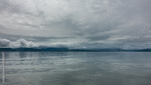 The ocean under a cloudy sky. Reflection on a shiny surface. In the distance, you can see a mountain range, the peaks of which are hidden in the clouds. Pastel shades. Petropavlovsk-Kamchatsky. 