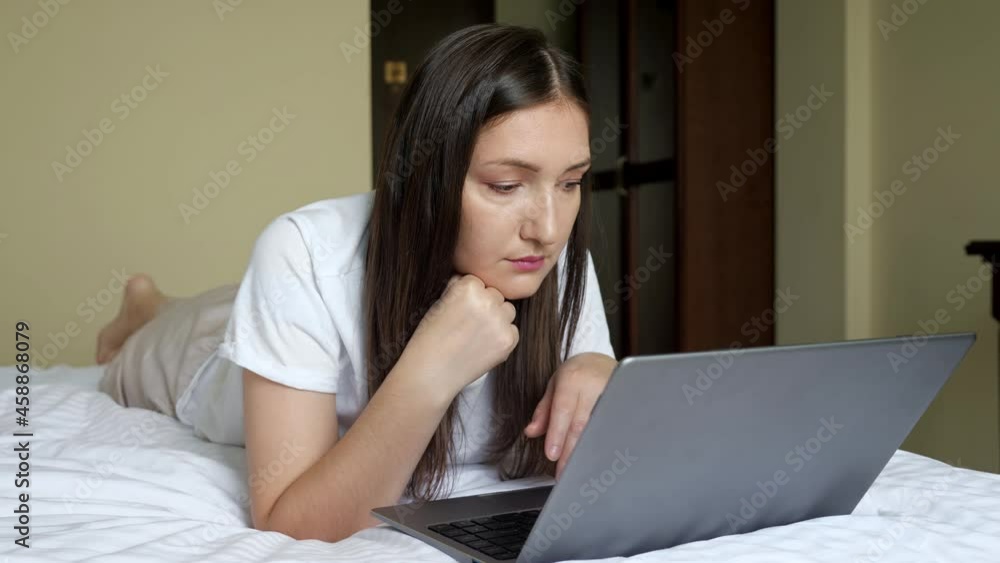 brunette woman working on laptop lying on stomach on bed. brunette woman working on laptop lying on stomach on bed.