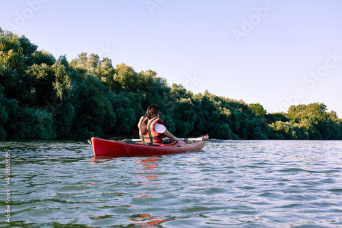 Rear view of woman rowing on red kayak on a Danube river in rural landscape with green trees at summer