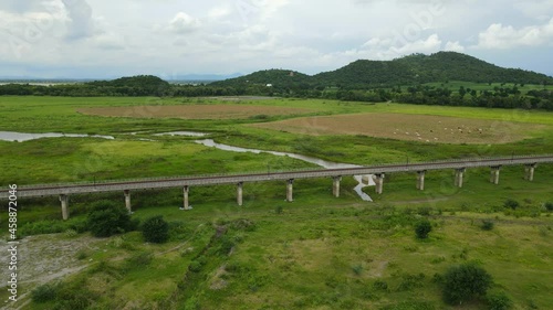 Angled aerial sliding footage towards the left revealing the elevated railways, farmland with cattle, mountains and lake in the horizon, Muak Klek, Saraburi, Thailand. photo