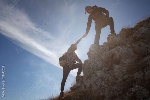 Silhouettes of two people climbing mountains and helping against the blue sky.
