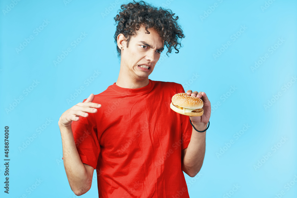 Cheerful guy with curly hair in a red t-shirt with a hamburger in his hands fast food diet Cheerful guy with curly hair in a red t-shirt with a hamburger in his hands fast food diet
