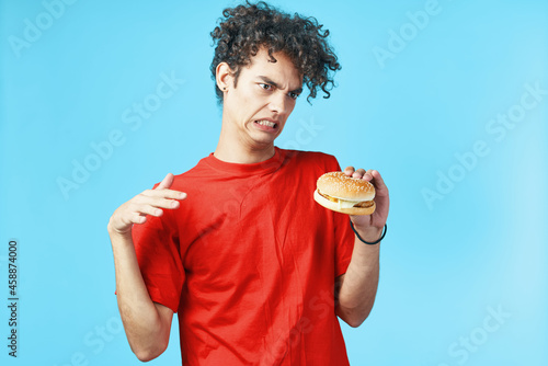 Cheerful guy with curly hair in a red t-shirt with a hamburger in his hands fast food diet