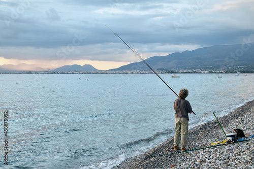 Fisherman with a fishing rod stands on the seashore against the backdrop of a beautiful sunset. Back view.