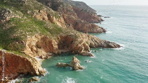 Summer landscape of rocky coastline of Portugal at Atlantic ocean. Aerial shot of ocean waves washing cliffs. Green grass growing on hills of mountains at sea on sunny day, 4k footage