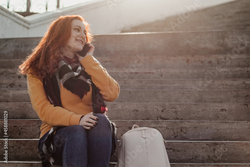 Young red-haired woman sitting on stairs and talking on phone. Lady in yellow sweater uses smartphone on sunny day. Lifestyle