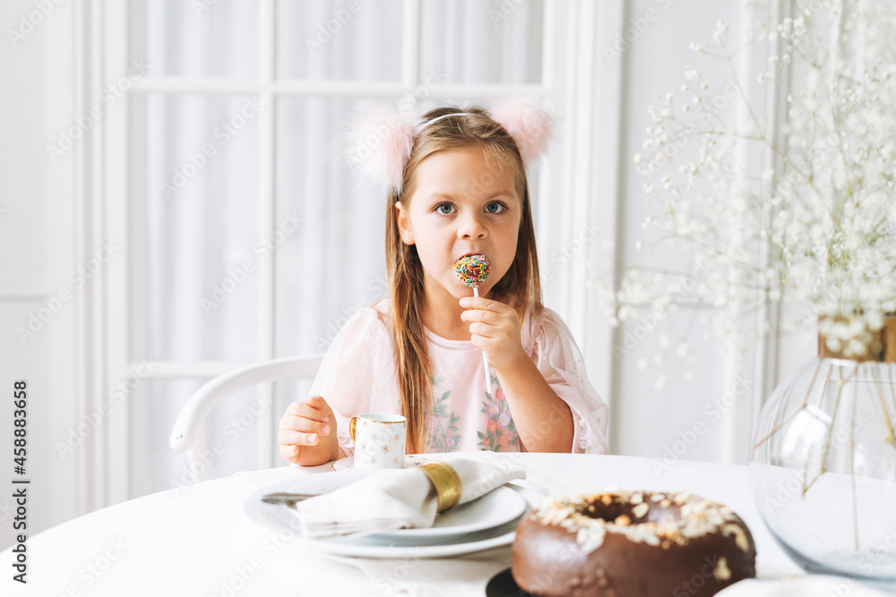 Funny cute little girl with long hair in light pink dress holding popcake in hands on festive table in bright living room at home. Christmas time, birthday girl Funny cute little girl with long hair in light pink dress holding popcake in hands on festive table in bright living room at home. Christmas time, birthday girl