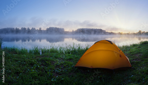 Orange tent on green grass of foggy lake at sunrise