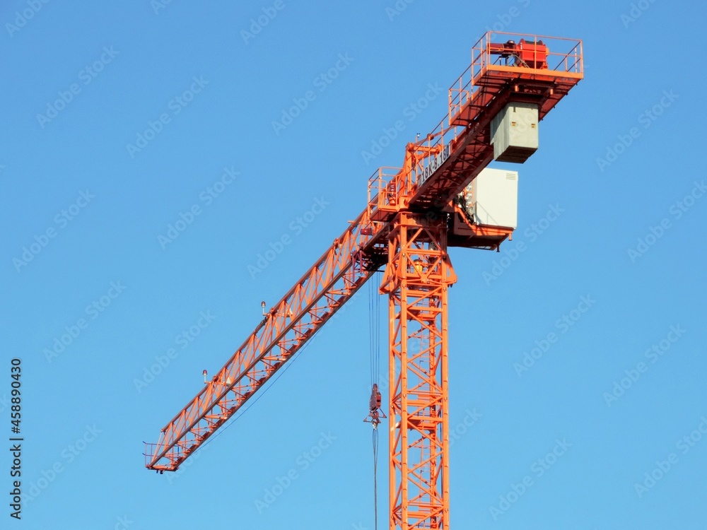 High-rise tower crane on the background of the blue sky. The tower crane is orange in color. High-rise tower crane on the background of the blue sky. The tower crane is orange in color.