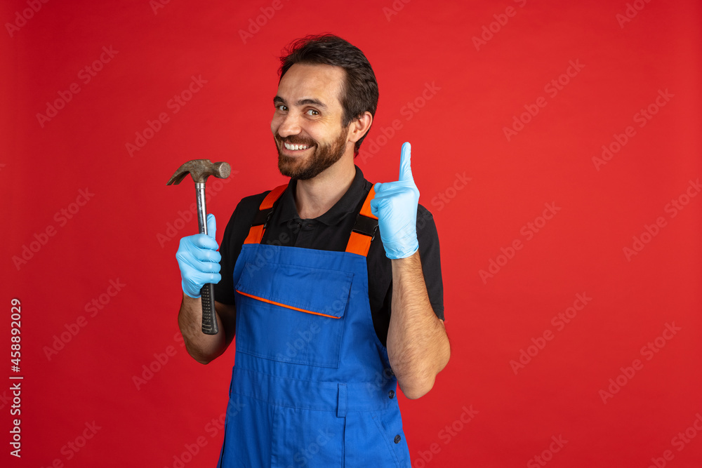 Funny young bearded man, male auto mechanic or fitter wearing blue work dungarees isolated over red studio background. Concept of facial emotions Funny young bearded man, male auto mechanic or fitter wearing blue work dungarees isolated over red studio background. Concept of facial emotions